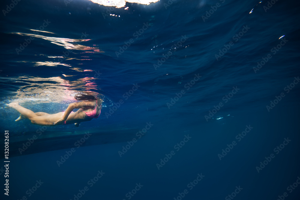 Girl in pink bikini underwater near bottom of a boat. Copy space on right side of image with blue water ocean background. Water surface with ripples above a model