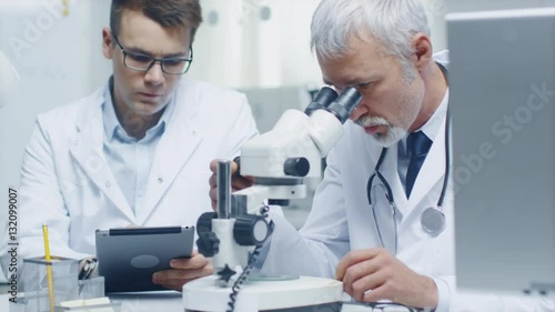 Senior Male Doctor Conducting Health Issue Investigation Through Microscope. His Young Assistant Writes all Relevant Data in Tablet Computer.Shot on RED Cinema Camera in 4K (UHD). photo