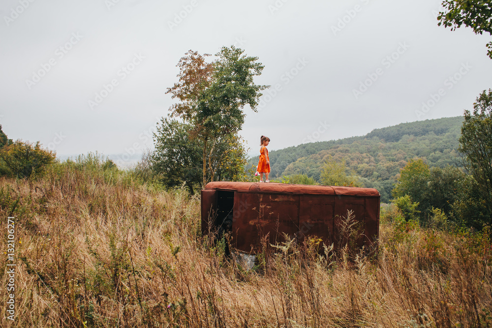 Naklejka premium little girl with a doll stands on the old trailer in the woods