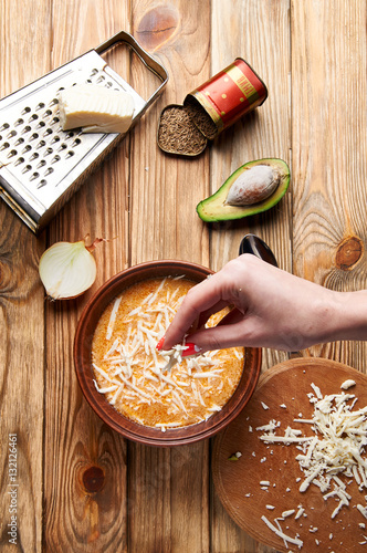 Female hand sprinkle with cheese delicious Argentinian cream soup. On the old wooden table lies a large grater, cheese, a plate of soup, sliced onions, avocado and cumin.