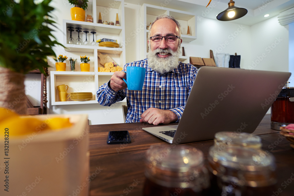 A pleased senior man holding a cup of tea while using laptop