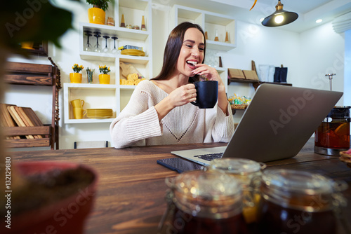 Laughing young woman working on laptop while holding a cup of te