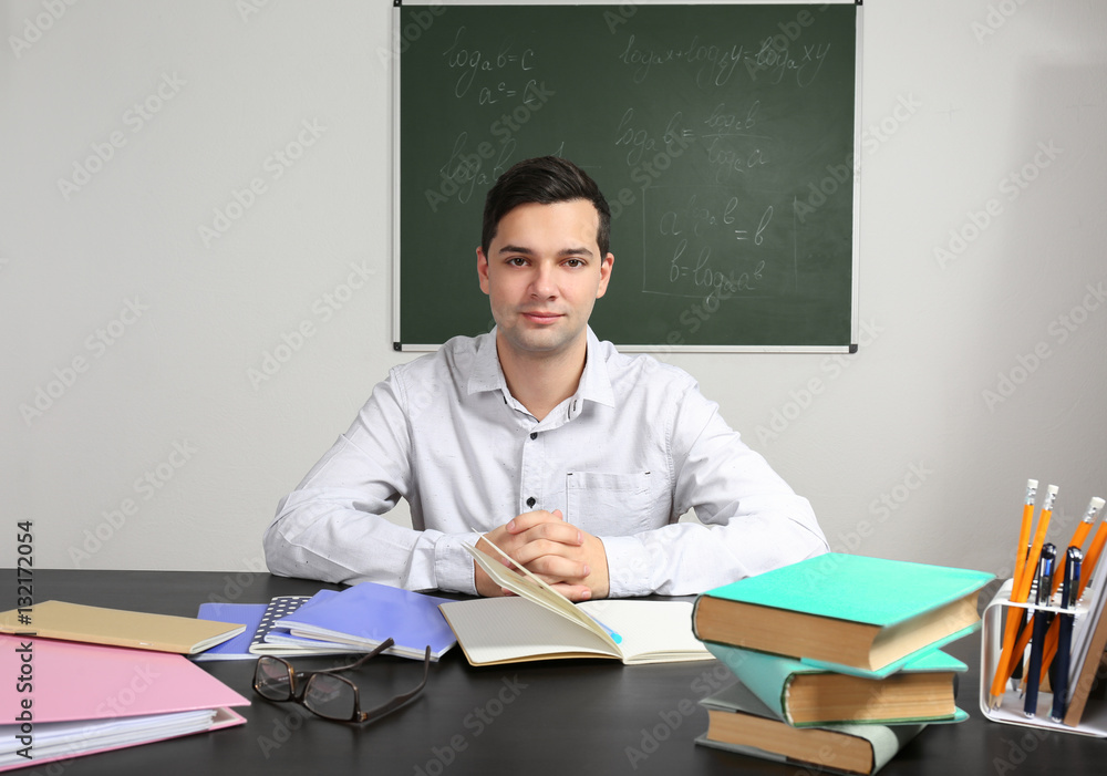 Handsome young teacher sitting at table in classroom Handsome young teacher sitting at table in classroom