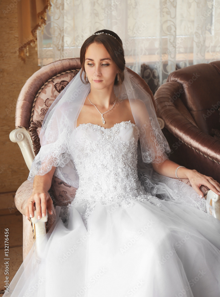 Fototapeta premium Elegant young bride in white wedding dress and veil sitting in ornate armchair, anticipating marriage ceremony
