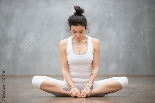 Portrait of beautiful young woman with floral tattoos working out against grey wall, doing yoga or pilates exercise, sitting in baddha konasana, bound angle, cobbler, butterfly pose. Full length shot