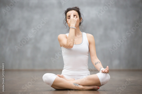 Front view portrait of beautiful young woman wearing white tank top working out against grey wall, resting after doing yoga exercises, using nadi shodhana pranayama technique. Full length