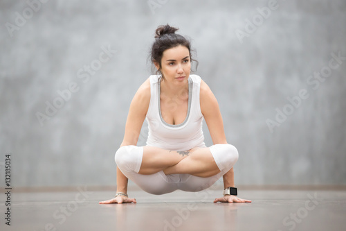 Portrait of beautiful young woman wearing white sportswear working out against grey wall, doing yoga or pilates exercise. Arm balance with crossed legs, Scale Posture, Tolasana, Utpluthi. Full length photo