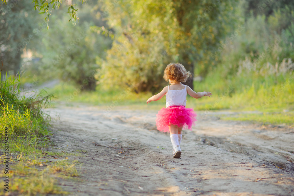 Little curly girl in a bright pink skirt tutu runs on a forest road, the view from the back. Little curly girl in a bright pink skirt tutu runs on a forest road, the view from the back.