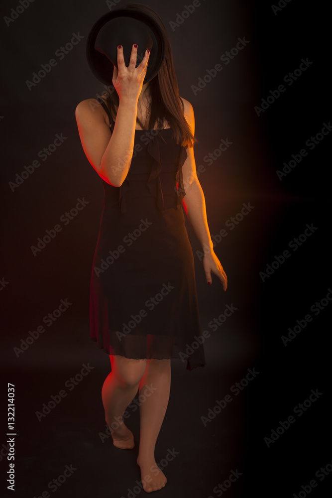 Beauty low key portrait of a young brunette woman with hat that cover face. Red backlight, black background