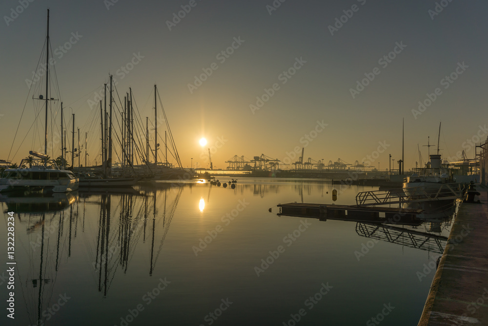 panoramic view sunrise at the Valencia harbor, the sun rises between docked sailboats and cargo port cranes, water reflection