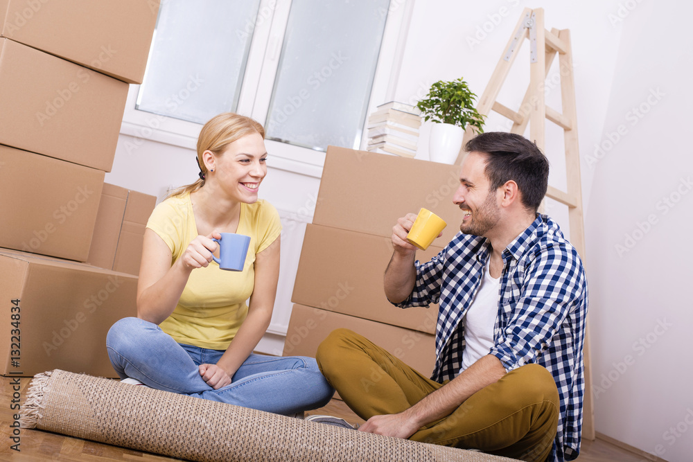 Young couple discussing plan of their new house and smiling while sitting on the floor among boxes and drinking coffee Young couple discussing plan of their new house and smiling while sitting on the floor among boxes and drinking coffee