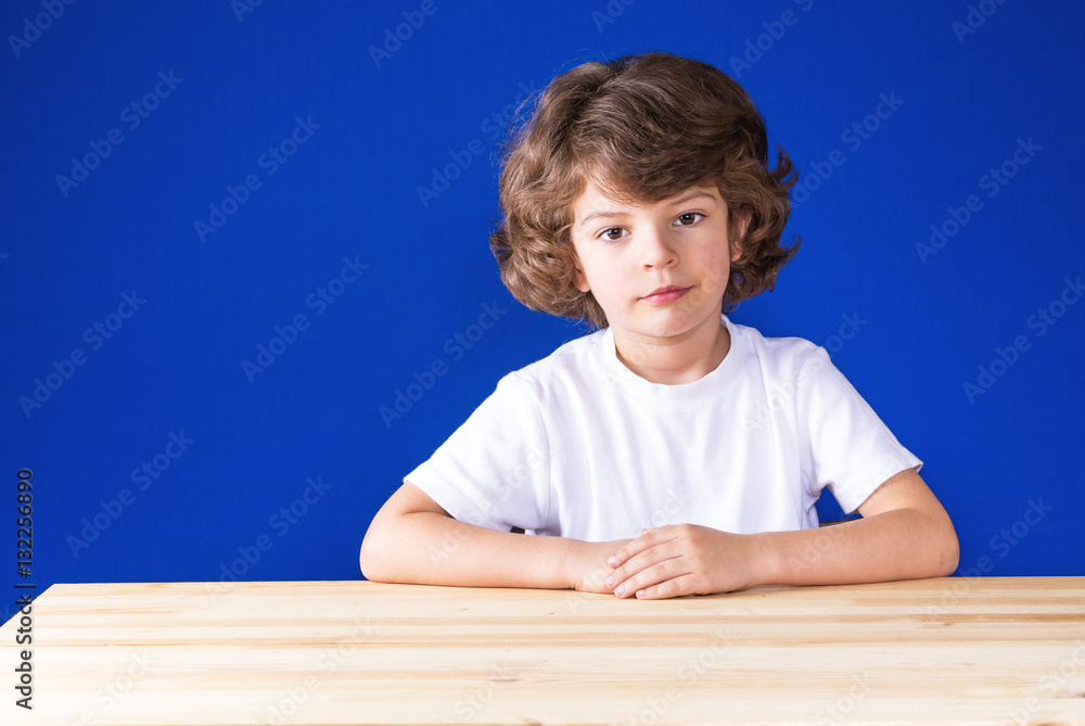 Serious curly cute boy sitting at a wooden table and sadly looks at the camera. Close-up. Blue background. Serious curly cute boy sitting at a wooden table and sadly looks at the camera. Close-up. Blue background.
