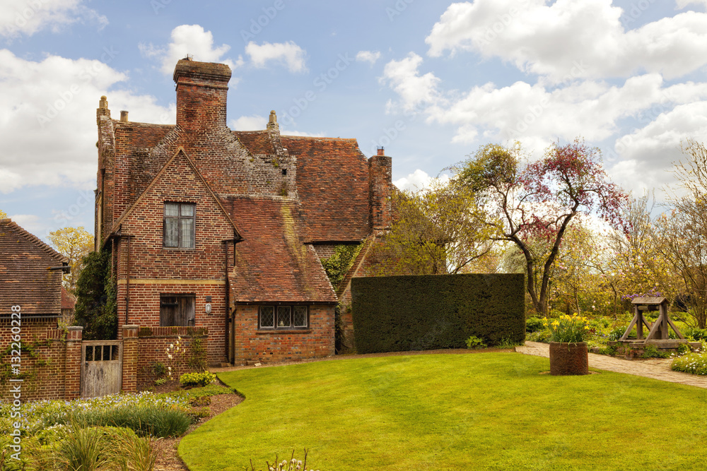 Old red brick english cottage in front of green lawn, with spring garden, water well