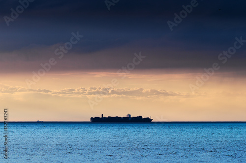 Silhouette of large container ship on the horizon crossing Baltic sea under dramatic dark nimbostratus cloud formation. Pomerania, Poland.