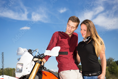 young couple looking sideways standing beside a motorcycle