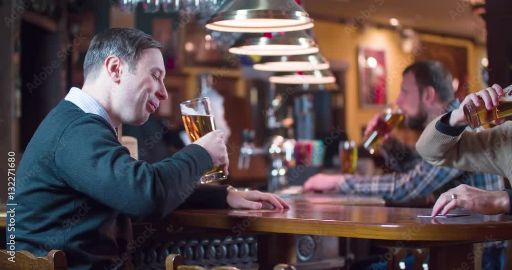 Men pub guests drinks beer at bar counter 4k video. Male friends doing toast, cheers, taste lager. Working bartender in background Men pub guests drinks beer at bar counter 4k video. Male friends doing toast, cheers, taste lager. Working bartender in background