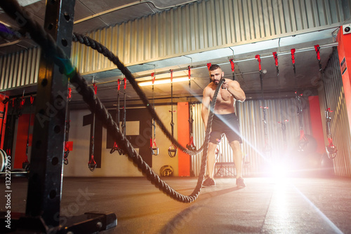 Young fit man exercising with battle ropes in the fitness gym