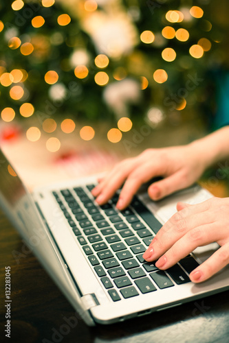 Last minute holiday shopping. Young woman shopping online on her laptop at Christmas time. Christmas tree lights out of focus in the background .