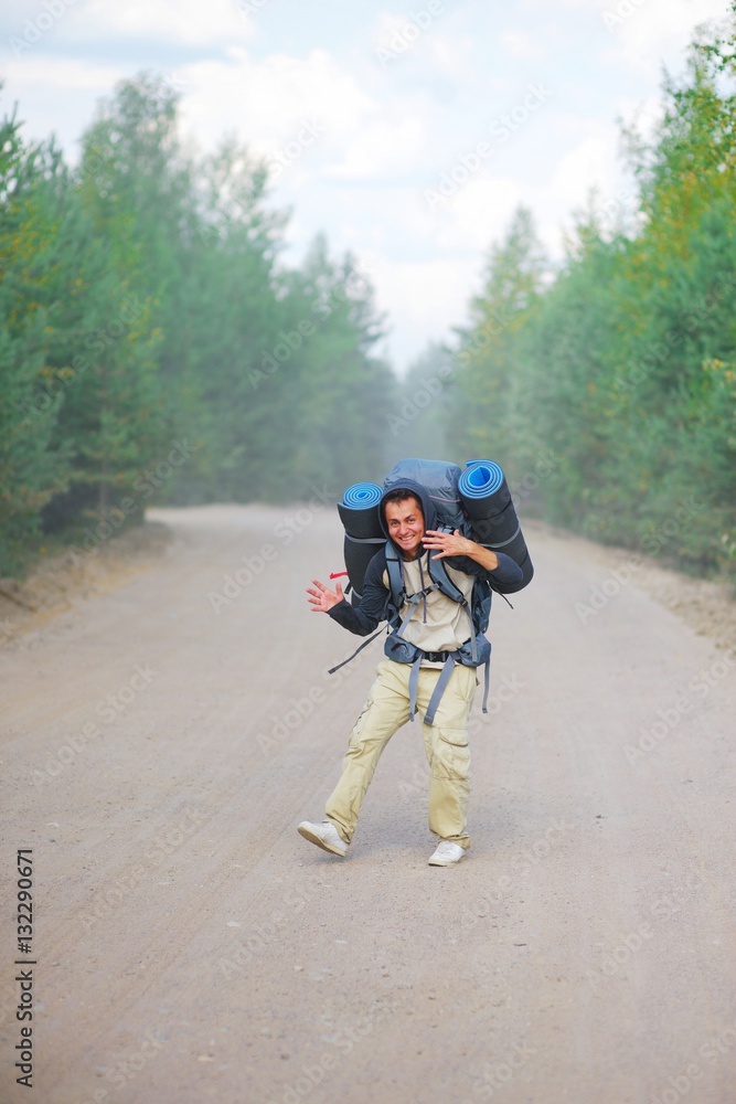 Fototapeta premium Young happy man in hoodie with heavy backpack and two travel rugs is on road through forest, waving his hands and laughing merrily. Hiking in the woods. Sport and concept of a healthy lifestyle.
