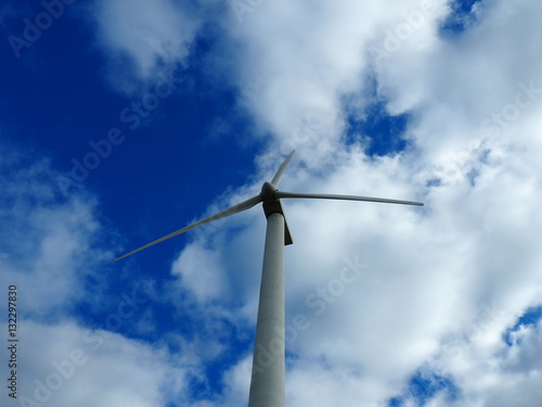 A wind turbine rises against a blue sky with scattered clouds. The tower and blades form a clear upward view of the structure from ground level.