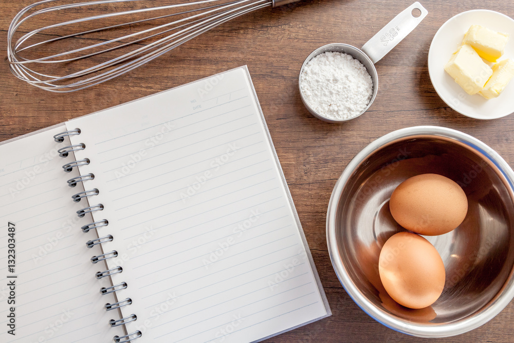 Blank notebook with eggs, butter, whisk and flour in measure cup on the wooden table