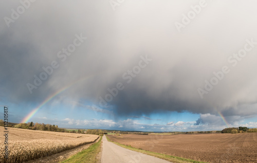 Panorama of the field by the road with a rainbow and stormy sky