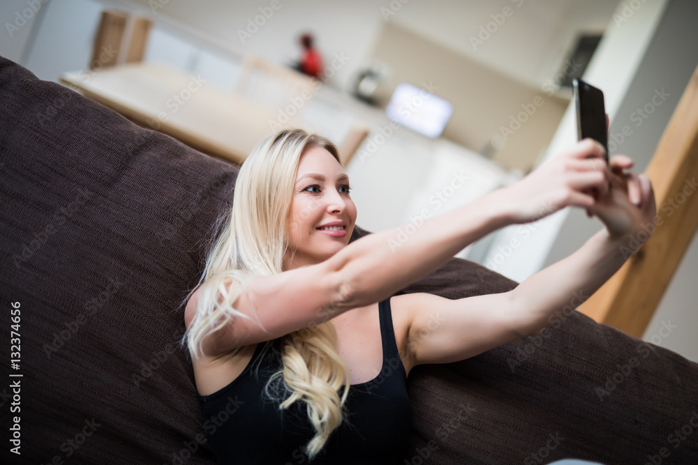 Fototapeta premium Blonde woman sitting on her sofa taking a picture of herself at home in the sitting room