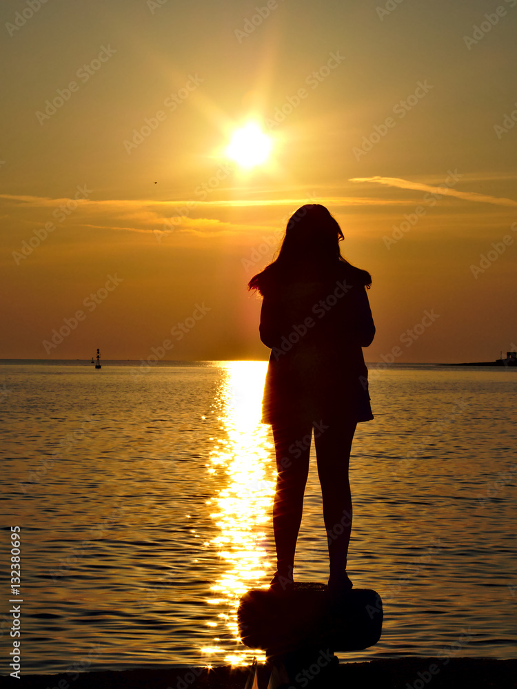 Silhouette of a woman standing on bollard at the harbor watching sea sunset..