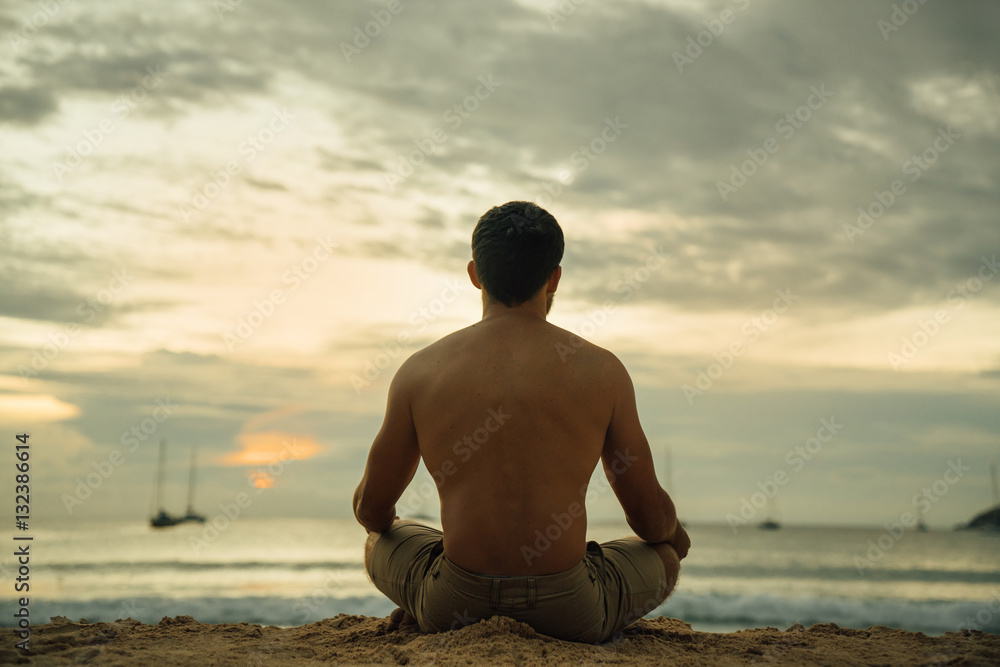 The guy looks at the sea sunset in a yoga pose. male fitness sitting on the beach practicing yoga. health and a clean mind.