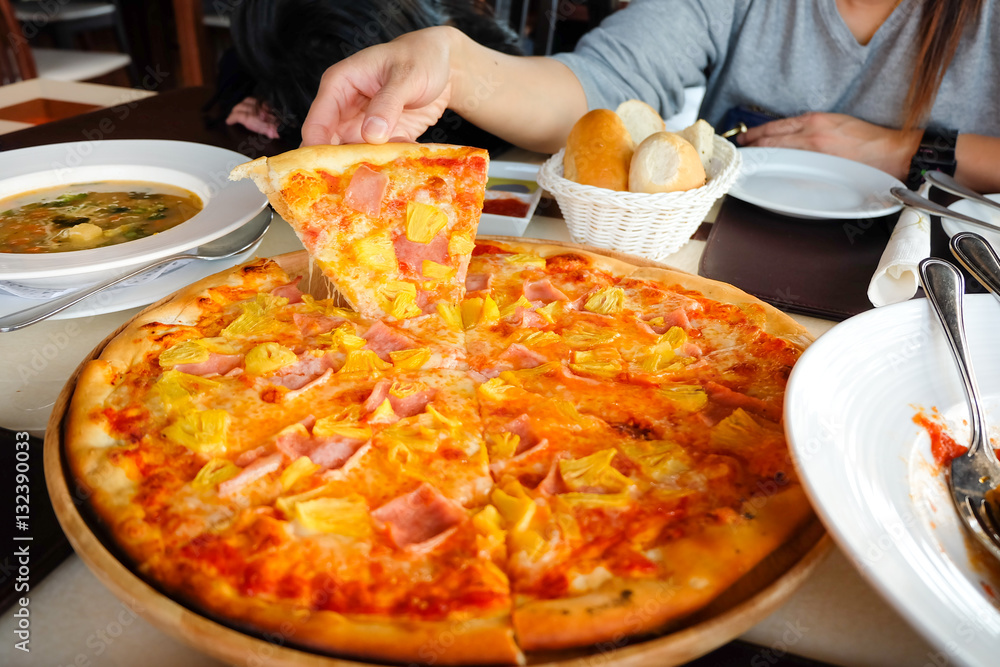 Woman's hand picked Hawaiian pizza from a wooden tray on the table.