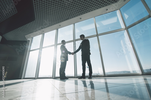 Two young businessmen are shaking hands with each other standing against panoramic windows. photo