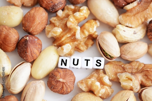 Raw mixed nuts and the word “nuts” spelled by tiled letter beads spread on a white table 