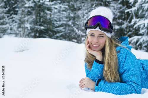 Smiling beautiful young woman with long blonde hair in snow jacket, white hat and goggles for skiing.