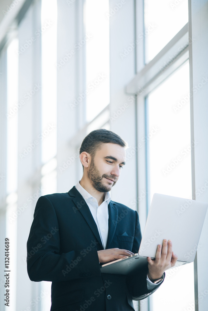 Handsome businessman in suit with laptop against windows in office