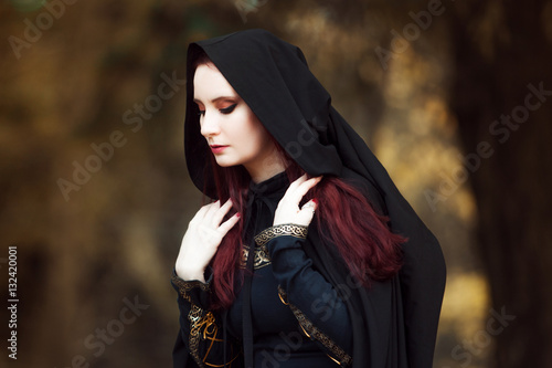 Young beautiful and mysterious woman in woods, in black cloak with hood, image of forest elf or witch © Ulia Koltyrina