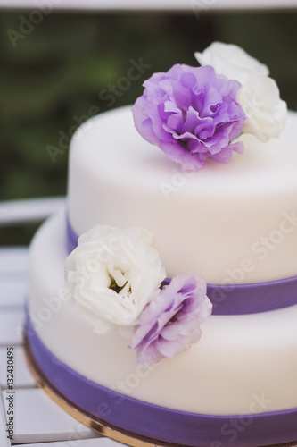 White wedding cake with purple flowers decoration outdoors on wooden modern chair.