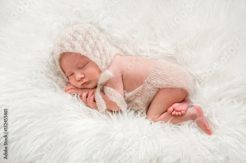 Новорожденный малыш в вязанном чепчике и боди спит на меховом пледе photo