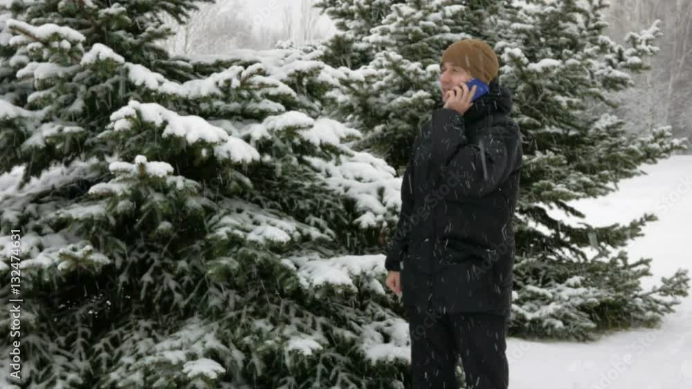A young man goes on a winter forest and talking on the phone. Big snowfall. He stops and ends the conversation. A man in a dark jacket and a warm hat. A young man goes on a winter forest and talking on the phone. Big snowfall. He stops and ends the conversation. A man in a dark jacket and a warm hat.