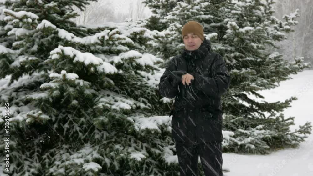 A young man in winter is cold in the forest near the spruce. Big snowfall. He's breathing on his hands, rubs and wears gloves with a hood. Snowy landscape. He looks into the camera A young man in winter is cold in the forest near the spruce. Big snowfall. He's breathing on his hands, rubs and wears gloves with a hood. Snowy landscape. He looks into the camera