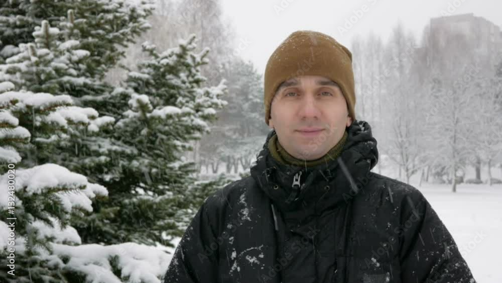 The young man looks into the camera and smiling in winter forest. Big snowfall. A man in a dark jacket and a warm hat. The young man looks into the camera and smiling in winter forest. Big snowfall. A man in a dark jacket and a warm hat.