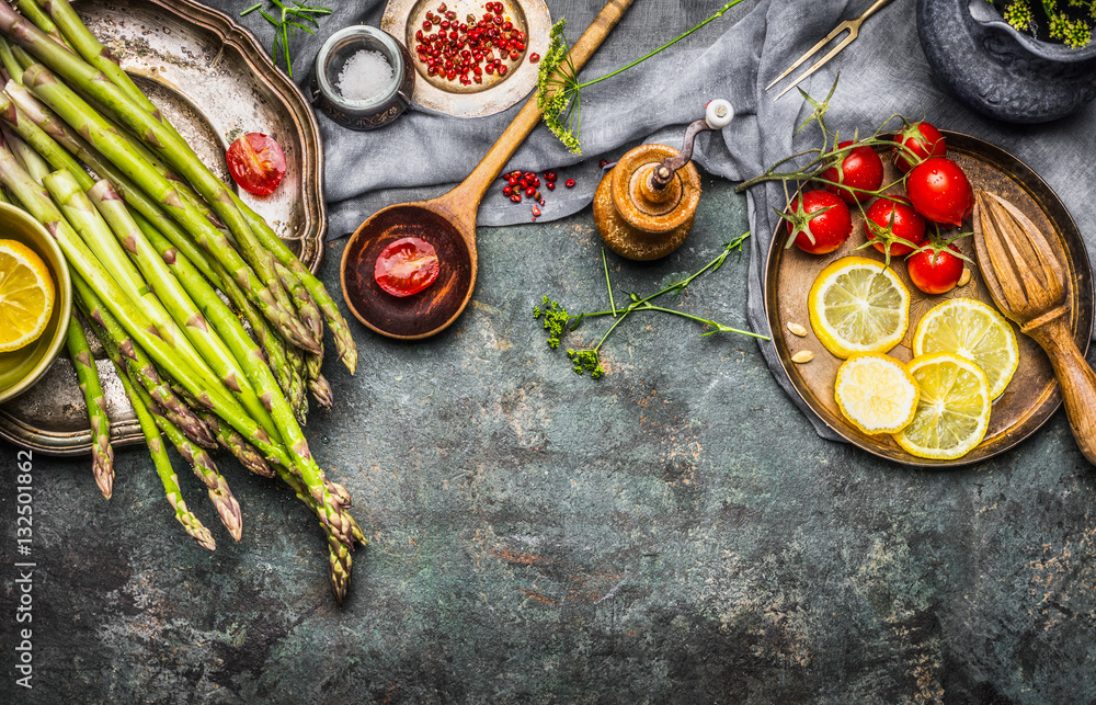 Tasty asparagus cooking with tomatoes, lemon and seasoning, preparation on rustic kitchen table with wooden spoon, top view, border. Dark style - obrazy, fototapety, plakaty 