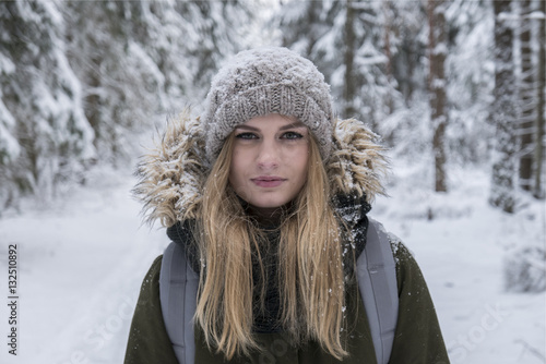 Young attractive Caucasian female hiker with blond hair smiling and looking into the camera, trees covered with snow in the background. Winter scenery, active lifestyle