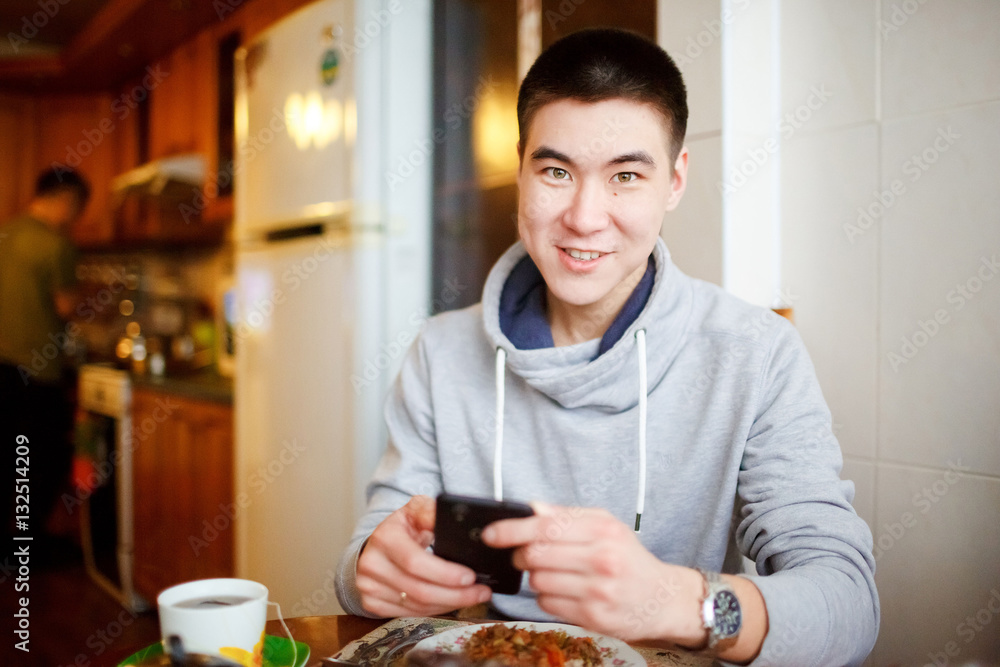 Positive sentiment of a young man sitting in the kitchen during breakfast holds in his hands the phone, looking at the camera. Positive sentiment of a young man sitting in the kitchen during breakfast holds in his hands the phone, looking at the camera.