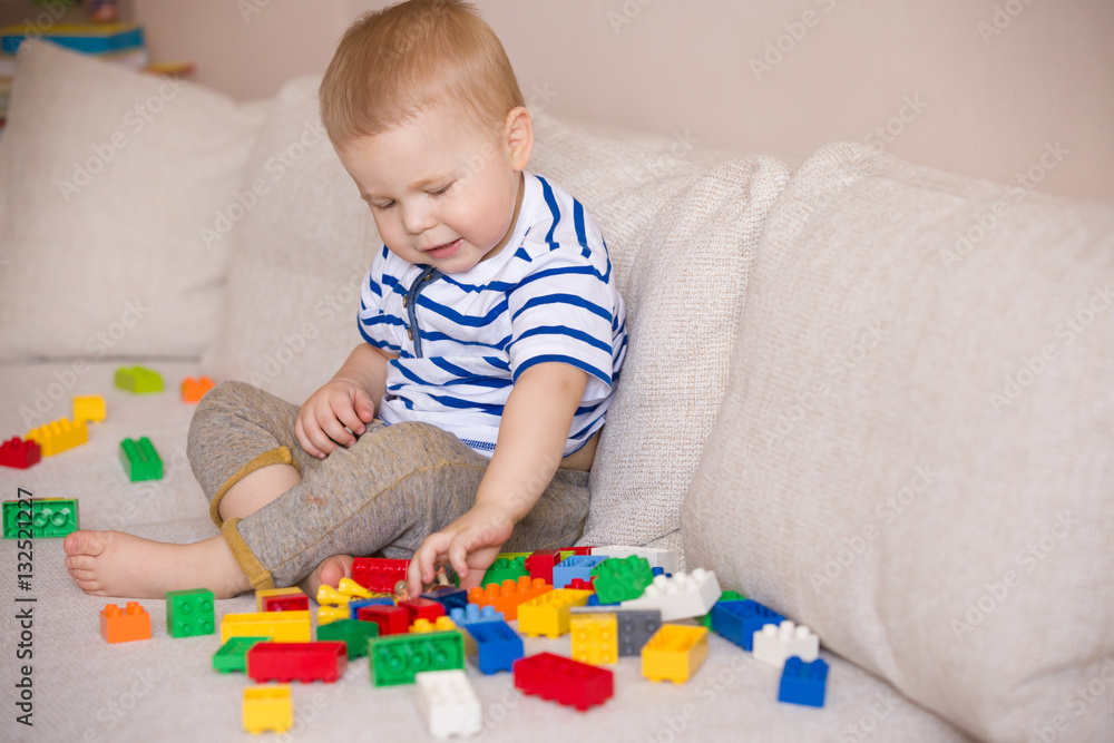 Cute little toddler boy in a striped shirt playing colorful plastic blocks on the sofa indoors. child having fun and building out of bright constructor bricks. Early learning. Creative.+ Cute little toddler boy in a striped shirt playing colorful plastic blocks on the sofa indoors. child having fun and building out of bright constructor bricks. Early learning. Creative.+