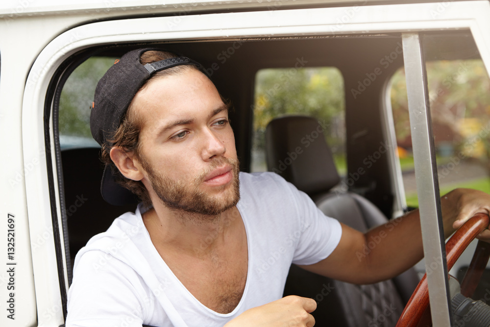 Attractive young Caucasian hipster man with beard wearing black baseball cap and white V-neck shirt driving white sport utility vehicle along country road, going to spend weekends in open air Attractive young Caucasian hipster man with beard wearing black baseball cap and white V-neck shirt driving white sport utility vehicle along country road, going to spend weekends in open air