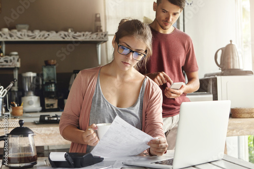 Young family facing financial troubles. Woman in glasses reading information in piece of paper with concentrated look while paying utility bills via internet using laptop computer and calculator photo