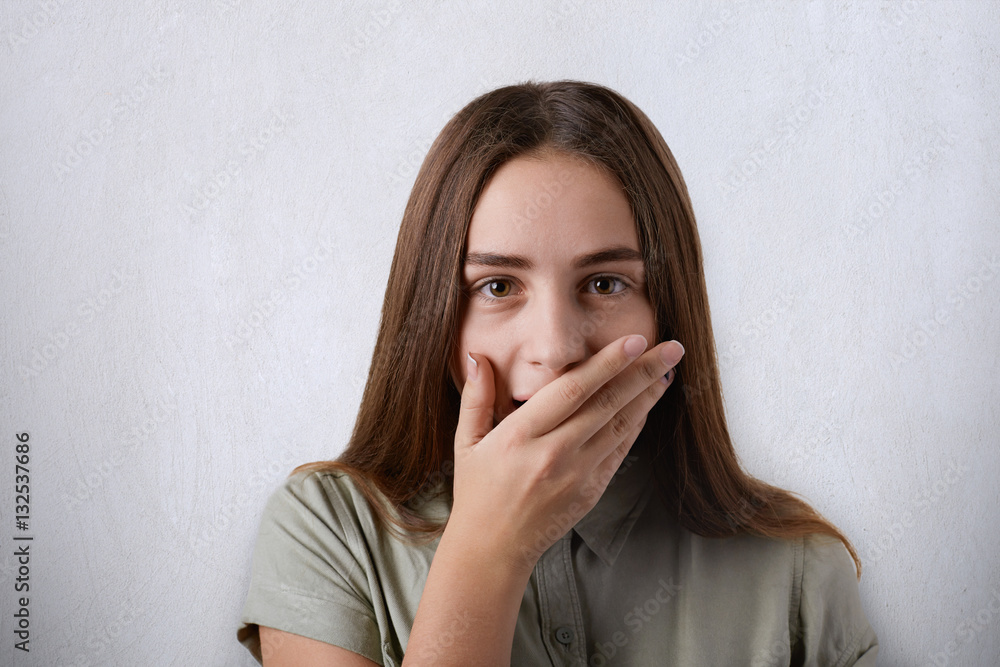 Surprised girl with long straight hair and dark eyes covering her mouth with hand over grey background and looking at camera. A surprised expression of young attractive woman. Shock condition Surprised girl with long straight hair and dark eyes covering her mouth with hand over grey background and looking at camera. A surprised expression of young attractive woman. Shock condition