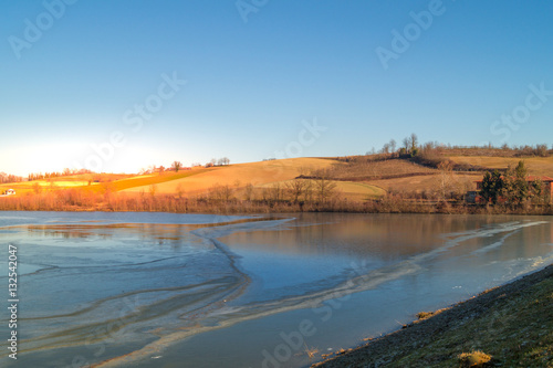 expanse of green grass on the lake with blue sky, countryside and farm around the lake, sunlit water