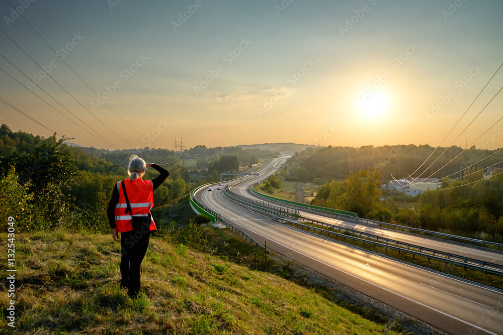 The man in an orange safety vest observing the highway with electronic toll system and the cars in the distance on the hill in the countryside at sunset. View from above.