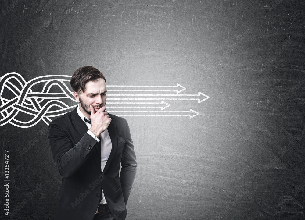 Bearded businessman in a suit is standing near a blackboard with tangled arrows sketch going straight. Mock up Bearded businessman in a suit is standing near a blackboard with tangled arrows sketch going straight. Mock up
